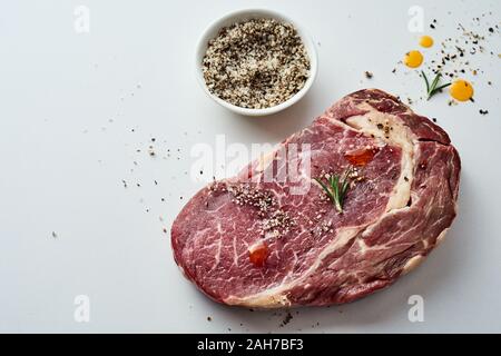 Porzione di grassi grezzi in marmo bistecca di manzo con sfregamento delle spezie e erbe su uno sfondo grigio con spazio copia vista elevato angolo Foto Stock