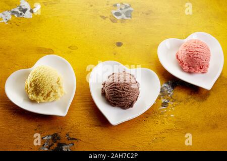 Cuore tre piatti sagomati con gelato porzioni in sapori diversi su un giallo sullo sfondo di legno Foto Stock