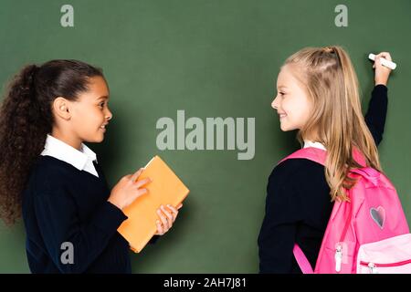 African American schoolgirl con libro in piedi accanto a un compagno di classe scrivere sulla lavagna verde Foto Stock