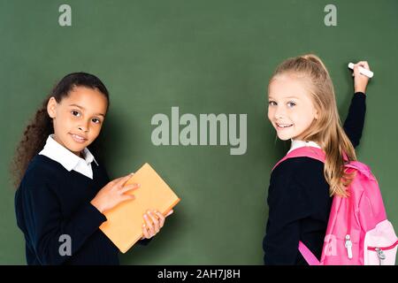 African American schoolgirl azienda prenota vicino al compagno di classe con un pezzo di gesso vicino a lavagna verde Foto Stock