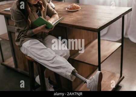 Vista ritagliata della ragazza con una gamba protesi libro lettura tabella accanto a casa Foto Stock