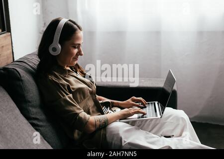 Vista laterale del freelancer sorridente nelle cuffie lavora al computer portatile sul divano Foto Stock