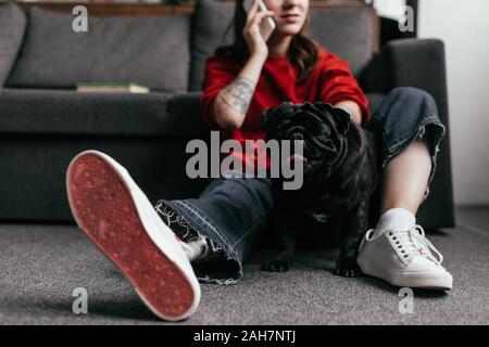 Vista ritagliata della donna con protesi di gamba parlando sullo smartphone accanto pug sul pavimento a casa Foto Stock