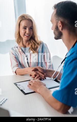 Messa a fuoco selettiva di donna sorridente stringono le mani con barbuto medico in clinica Foto Stock