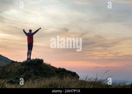 Sagoma scura di un escursionista scalare una montagna al tramonto alzando le mani in piedi sulla vetta come un vincitore. Foto Stock