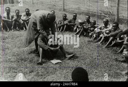 Kenya, un Mau Mau detenuto seduto al centro di un cerchio di uomini in un recintato composto è purificate dei Mau Mau giuramento.manoscritto originale didascalia: De-oathing, 1953. 1995/076/1/1/15/3.18. Foto Stock