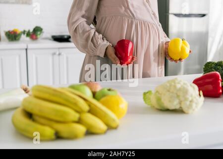 Vista ritagliata della donna incinta tenendo la paprica in piedi vicino a frutti sul tavolo Foto Stock