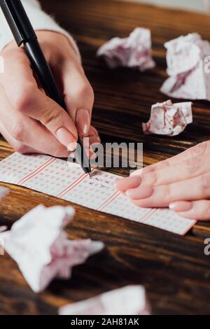 Vista parziale del giocatore i numeri di marcatura nel biglietto della lotteria vicino accartocciata lotteria carte sul tavolo di legno Foto Stock