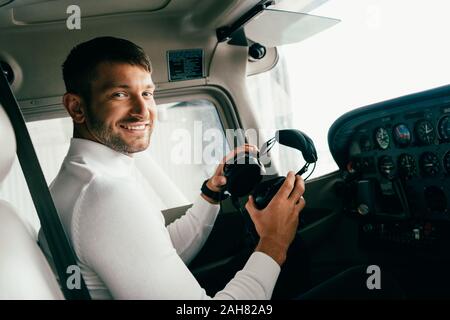 Sorridente barbuto giovane uomo in abbigliamento casual partecipazione delle cuffie in aereo Foto Stock