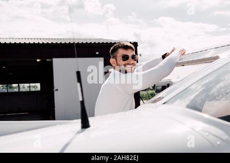 Sorridente uomo barbuto in occhiali da sole in piedi vicino a piano nella giornata di sole Foto Stock