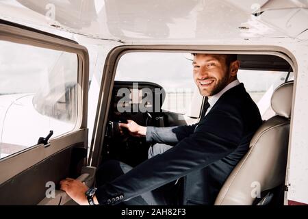 Pilota sorridente in abbigliamento formale seduto in aereo e holding Foto Stock