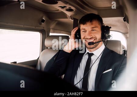 Sorridente pilota barbuto in abbigliamento formale e la cuffia seduto in aereo Foto Stock