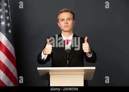 Kiev, Ucraina - 18 ottobre 2019: emotional man imitando Donald Trump mostrando pollice in alto sulle tribune con bandiera americana su sfondo nero Foto Stock