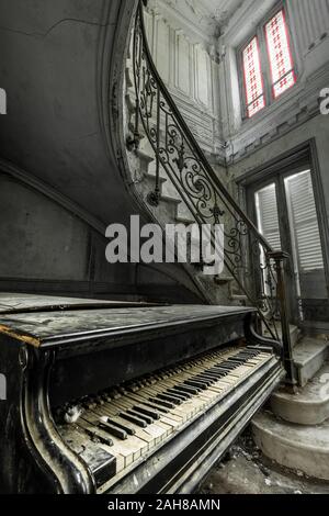 Vista ravvicinata di un antico pianoforte a coda stagionato adagiato lungo la scala della scala a chiocciola in una casetta francese abbandonata Foto Stock