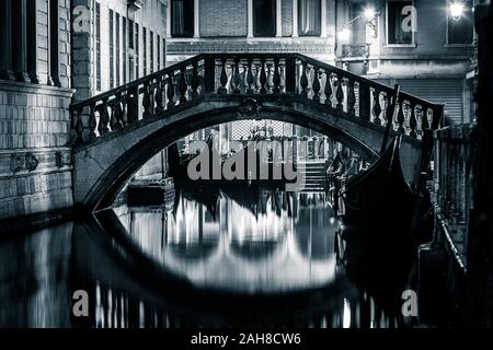 Iconico scatto notturno in bianco e nero di un canale veneziano con gondole a galla e un ponte di marmo che riflette sull'acqua Foto Stock