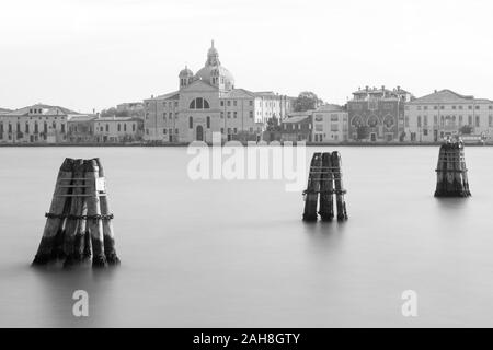 Paesaggio urbano di Venezia, con tre pali ormanti che emergono dall'acqua in primo piano, e un'antica chiesa sullo sfondo Foto Stock