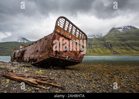 Vista grandangolare di una vecchia nave da carico arrugginita e relegata, bloccata in un fiordo, con montagne lontane sotto un cielo nuvoloso scuro Foto Stock