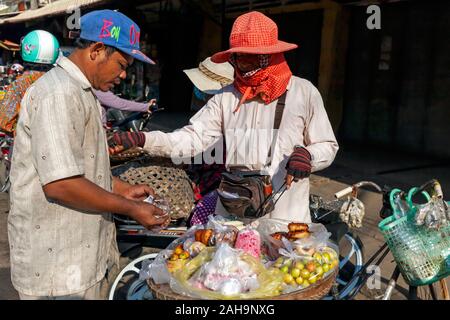 Un uomo asiatico è di acquistare il cibo da una donna per la vendita di frutta, deserto e ciambelle legato alla sua bicicletta ad un mercato di strada in Kampong Cham, Cambogia. Foto Stock