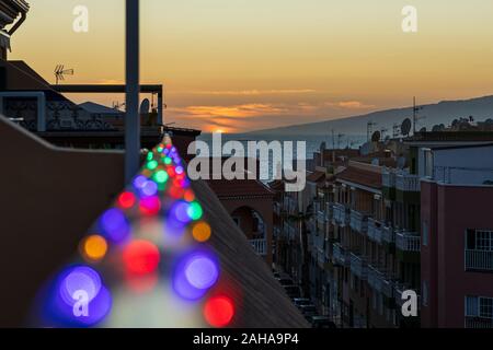 Impostazione di Sun in mare alla vigilia di Natale con le luci di Natale al di fuori della messa a fuoco in primo piano, visto da un alto punto di vantaggio, Playa San Juan, Tenerife, Foto Stock