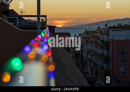 Impostazione di Sun in mare alla vigilia di Natale con le luci di Natale al di fuori della messa a fuoco in primo piano, visto da un alto punto di vantaggio, Playa San Juan, Tenerife, Foto Stock