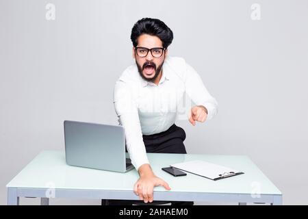 Arrabbiato uomo barbuto in tuta in piedi appoggiato le mani sulla scrivania con computer portatile e grida di office Foto Stock