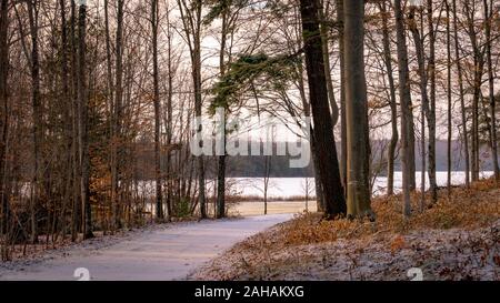 Una strada coperta di neve si curva verso un lago ghiacciato in cottage paese durante l'inverno. Alberi circondano. Foto Stock