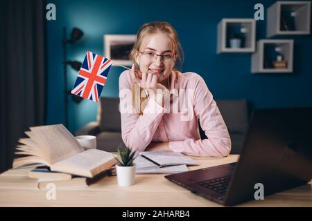 Lo zenzero carina ragazza con gli occhiali seduto alla scrivania e appoggiando il suo mento tenendo una bandiera britannica Foto Stock