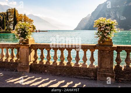 Lago di Garda promenade con la classica pietra ringhiere di recinzione costruito sul bordo con vasi da fiori con rigogliosi fiori bianchi. Lago di Garda circondato Foto Stock