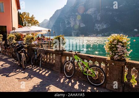 Riva del Garda, Lombardia, Italia - 12 Settembre 2019: Biciclette parcheggiate vicino al recinto di pietra ringhiere vicino al ristorante all'aperto pieno di persone su Gard Foto Stock