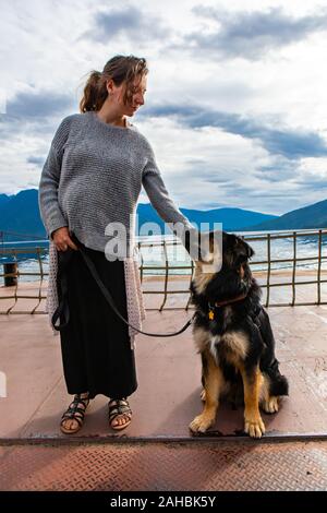 Giovane donna si erge con la sua seduta cane nero al guinzaglio mentre prendere un traghetto con una vista panoramica delle montagne dietro Foto Stock