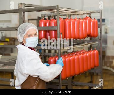 Lavoratore di sesso femminile che trasportano materie salsicce in rack in stanza di deposito alla trasformazione della carne factory Foto Stock