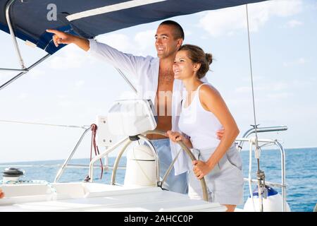 Uomo sorridente che punta a qualcosa su ocean orizzonte a sua moglie mentre in piedi sulla ruota di yacht godendo Estate Mare Vacanze Foto Stock