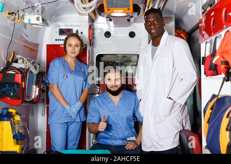 Tre positivi sorridente cordiale efficiente ambulanza medici che pongono in ambulanza con attrezzature mediche Foto Stock