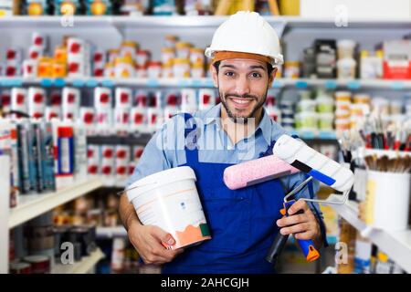 Soddisfatto giovane maestro in piedi tra le scaffalature in magazzino vernici con rulli e vernice Foto Stock