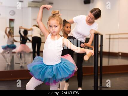 Giovani femmine di danza classica con insegnante aiutando il suo piccolo studenti vicino a ballet barre in sala da ballo Foto Stock