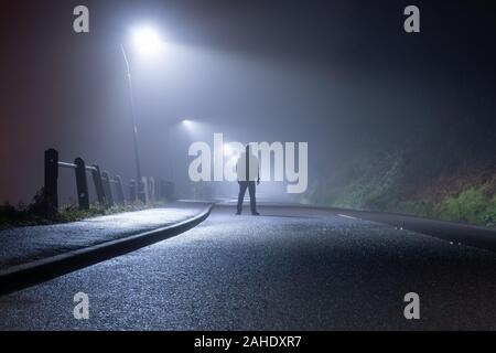 Un uomo misterioso, da soli, in piedi nel mezzo di una strada di campagna. Sotto le luci di strada. In una nebbiosa, Moody, Spooky, inverni di notte Foto Stock