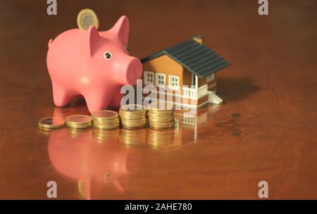 Un salvadanaio con una piccola casa e alcune monete su un tavolo di legno con riflessioni. Immagine concettuale circa il risparmio per l'acquisto di una casa. Foto Stock
