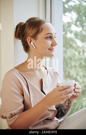 Attraente sorridente giovane donna di bere il caffè e ascoltare della buona musica quando si è seduti sul davanzale Foto Stock