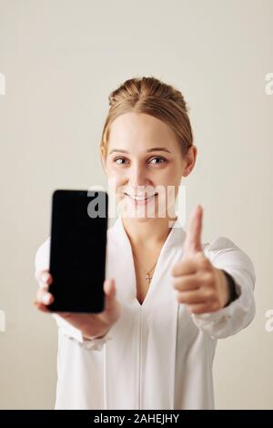 Ritratto di eccitato giovane donna che mostra smartphone con vuoto nella schermata nera e il pollice in su Foto Stock