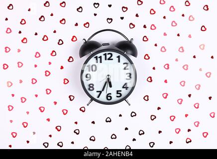 Nero orologio sveglia su uno sfondo bianco di sequin cuori Foto Stock