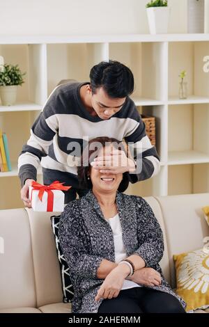 Giovane uomo asiatico che copre gli occhi di sua madre e dando il suo presente sulle madri giorno Foto Stock