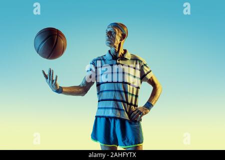 Senior uomo che indossa sportwear giocare a basket sul gradiente dello sfondo, una luce al neon. Caucasica modello maschile in grande forma rimane attivo. Concetto di sport, attività, movimento, benessere e fiducia. Foto Stock