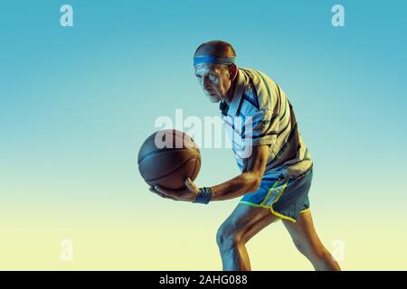 Senior uomo che indossa sportwear giocare a basket sul gradiente dello sfondo, una luce al neon. Caucasica modello maschile in grande forma rimane attivo. Concetto di sport, attività, movimento, benessere e fiducia. Foto Stock