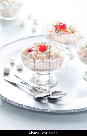 Una vista ravvicinata di un piatto da dessert riempito con insalata di ambrosia e rabboccato con ciliegie al maraschino su un metallo che serve il vassoio contro una finestra piena di sole. Foto Stock