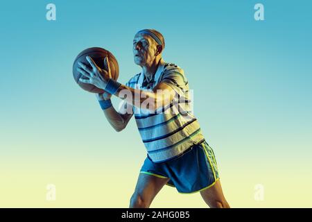 Senior uomo che indossa sportwear giocare a basket sul gradiente dello sfondo, una luce al neon. Caucasica modello maschile in grande forma rimane attivo. Concetto di sport, attività, movimento, benessere e fiducia. Foto Stock