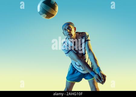 Senior uomo che indossa sportwear giocando a pallavolo sul gradiente dello sfondo, una luce al neon. Caucasica modello maschile in grande forma rimane attivo. Concetto di sport, attività, movimento, benessere e fiducia. Foto Stock