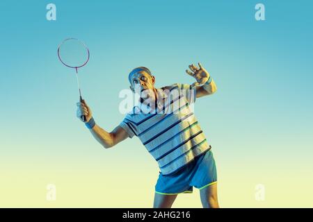 Senior uomo che indossa sportwear giocando badminton sul gradiente dello sfondo, una luce al neon. Caucasica modello maschile in grande forma rimane attivo. Concetto di sport, attività, movimento, benessere e fiducia. Foto Stock