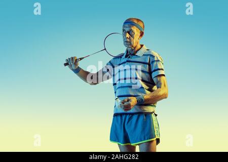 Senior uomo che indossa sportwear giocando badminton sul gradiente dello sfondo, una luce al neon. Caucasica modello maschile in grande forma rimane attivo. Concetto di sport, attività, movimento, benessere e fiducia. Foto Stock