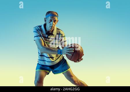 Senior uomo che indossa sportwear giocare a basket sul gradiente dello sfondo, una luce al neon. Caucasica modello maschile in grande forma rimane attivo. Concetto di sport, attività, movimento, benessere e fiducia. Foto Stock