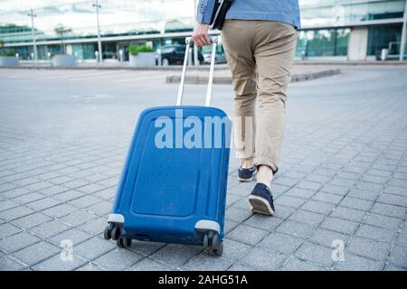 Uomo elegante a piedi per l'aeroporto con valigia blu. Uomo d'affari in viaggio. Foto Stock